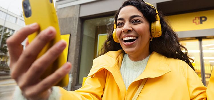 Junge Frau mit gelbem Smartphone und gelber Jacke lacht und macht ein Selfie